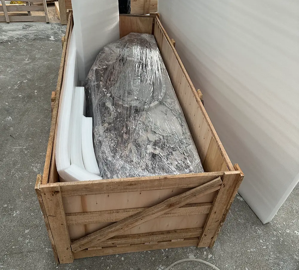 Top-down view of a custom marble sink being packaged in a factory setting. The heavy stone vanity, featuring an integrated basin, is tightly wrapped in clear protective plastic film and sits inside a sturdy, custom-built wooden export crate. Thick white high-density foam padding strips are positioned alongside the stone to absorb shock. The crate rests on a concrete workshop floor, ready for international shipping.