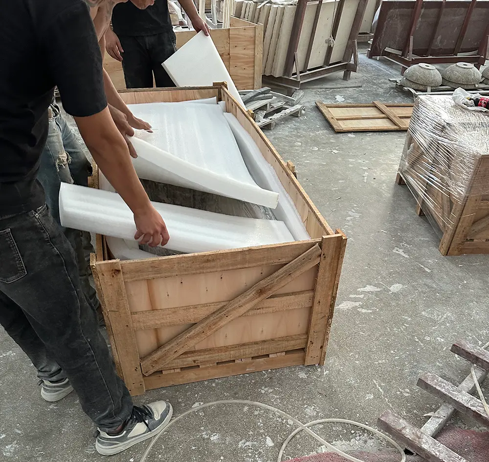 Custom Marble Sink Secure Factory Packaging & Export Crating Two factory workers in a stone fabrication workshop carefully placing thick white high-density foam padding over a shrink-wrapped custom marble sink. The heavy stone vanity sits inside a reinforced, custom-built wooden export crate on a concrete floor. Other wooden shipping crates and carved stone products are visible in the background, highlighting the factory's hands-on global logistics and packaging process.