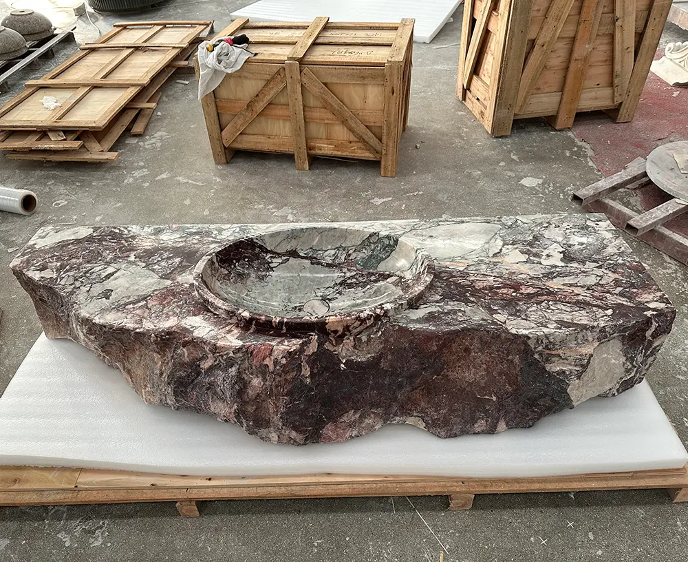 Front view of a massive custom natural marble sink resting on thick white protective foam and a wooden shipping pallet on a concrete factory floor. The sink has a polished top, a shallow integrated circular washbasin, and a heavily chiseled raw rock front edge. The stone is a breccia marble featuring dark burgundy, grey, white, and subtle green veining. Sturdy wooden export shipping crates and workshop materials are visible in the background, highlighting the stone packaging process.