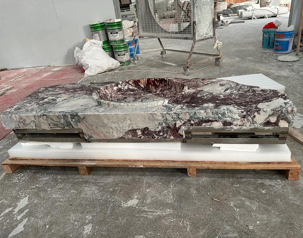 Custom Raw Edge Marble Sink Factory Fabrication & Mounting System14 Front-angle view of a heavy custom natural marble sink resting on protective white foam and a wooden shipping pallet inside a stone fabrication workshop. The breccia marble features deep burgundy, white, and subtle green veining. It has a polished top, an integrated circular basin, and a heavily chiseled raw rock front edge. A heavy-duty steel mounting frame is attached underneath the stone for wall-hung installation. Industrial workshop equipment, including a large fan and chemical barrels, is visible in the background.