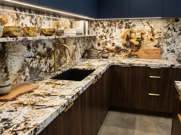 A modern luxury kitchen featuring Patagonia Quartzite countertops, a continuous full-height backsplash, and a floating stone shelf. The exotic natural stone displays dramatic white translucent quartz, deep black spots, and golden-brown veining. The kitchen design includes dark walnut lower cabinets with gold pulls, matte navy upper cabinets with under-cabinet LED lighting, a black undermount sink, and a brushed brass faucet. The counters are styled with rustic ceramic bowls and wooden cutting boards.