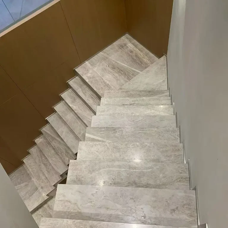 Top-down view of a modern indoor staircase clad in polished Taj Mahal Quartzite with natural beige and grey veining, featuring metal edge trims, bordered by a wood panel wall on the left and a white wall on the right.