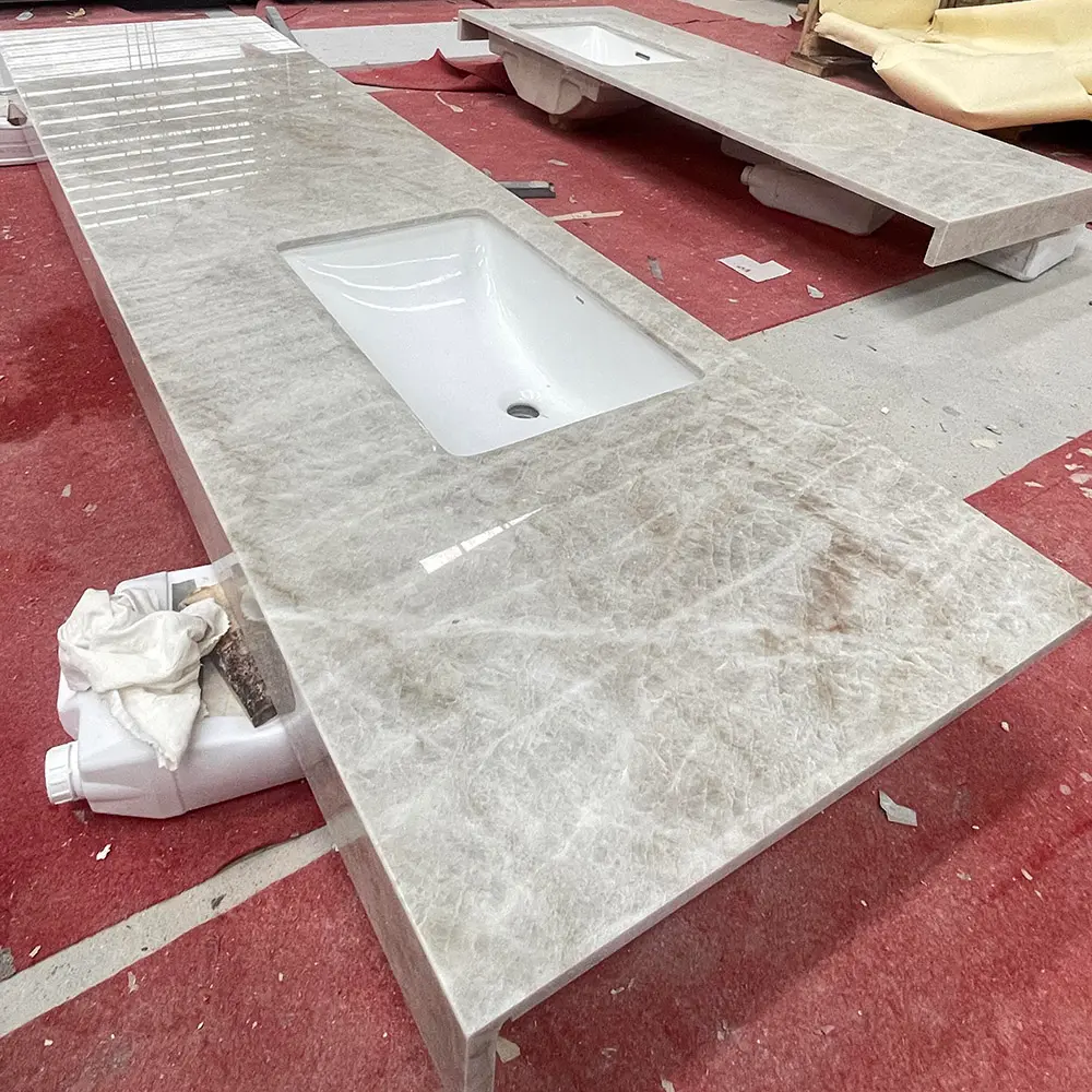 Taj Mahal Quartzite Extended Vanity Top with Integrated Sink and Mitered Apron Long highly polished Taj Mahal Quartzite bathroom vanity top with an integrated rectangular white undermount sink and mitered apron edge, staged on a red protective floor mat inside a stone fabrication factory.