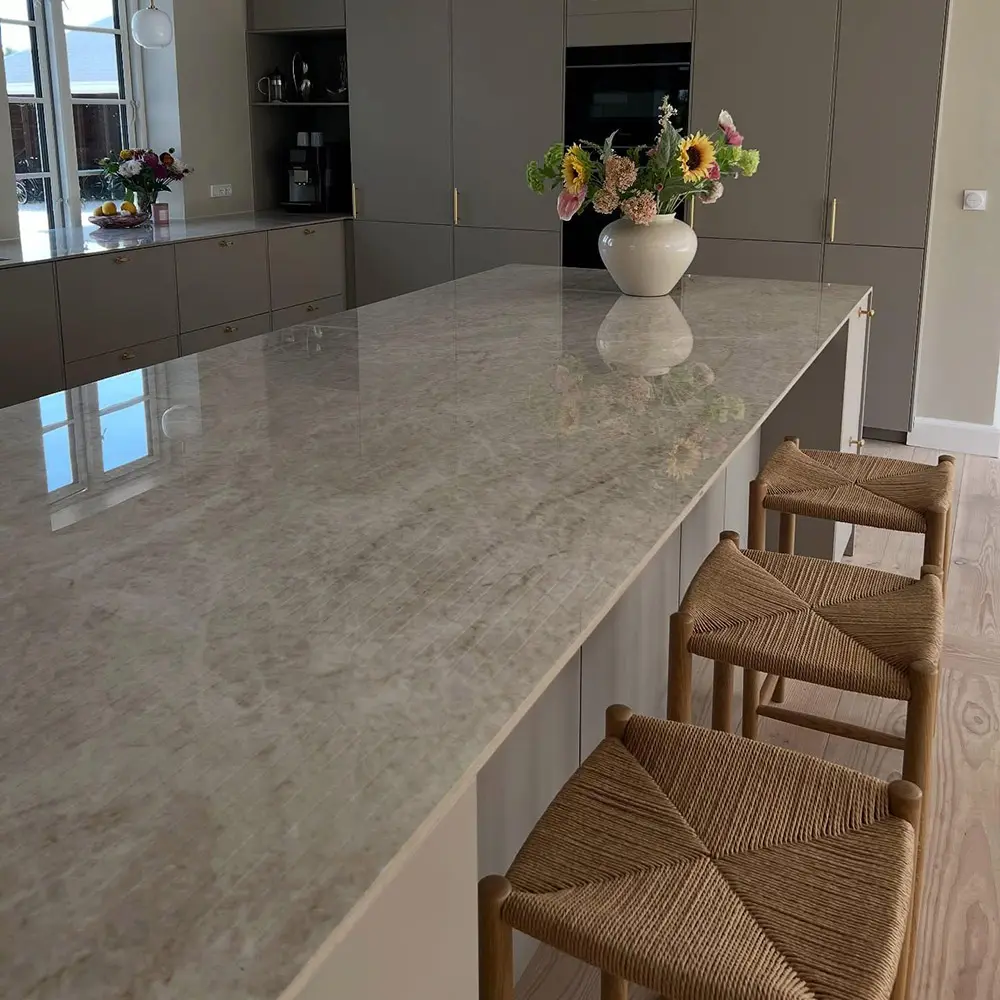 Taj Mahal Quartzite-Seamless Kitchen Island & Cabinet Integration Highly polished Taj Mahal Quartzite kitchen island countertop reflecting natural window light, integrated with flat-panel taupe cabinets and paired with three woven wooden bar stools.