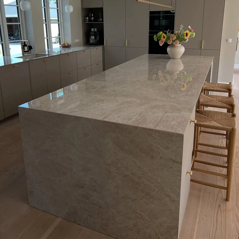 Large Taj Mahal Quartzite kitchen island with a seamless mitered waterfall edge dropping vertically to a light wood floor, reflecting natural light in a modern architectural kitchen.