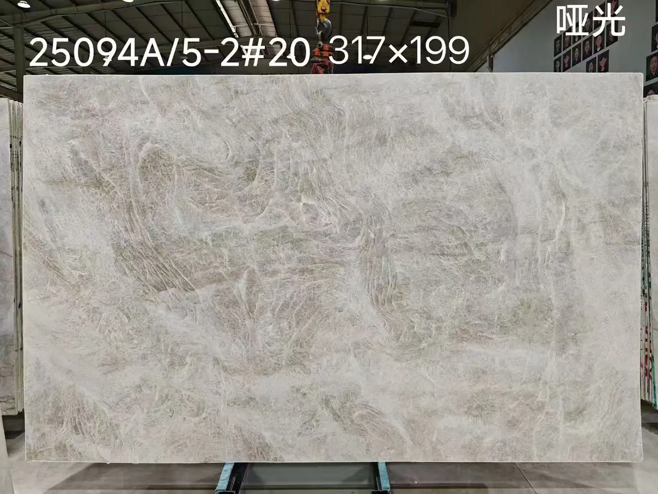 honed-taj-mahal-quartzite-slab Large honed Taj Mahal Quartzite jumbo slab measuring 317 by 199 cm with factory lot numbers, showing complex grey and beige veining, suspended by a crane in an industrial warehouse.