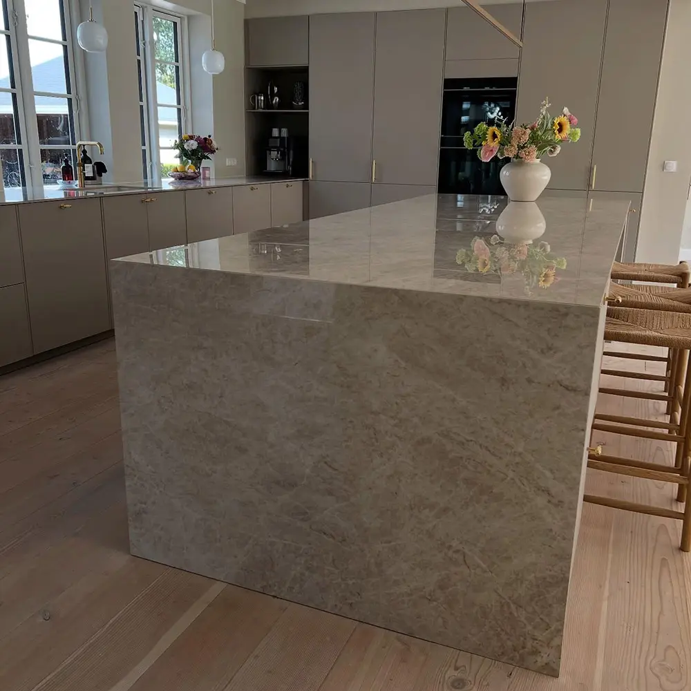 taj-mahal-quartzite-waterfall-island-caron-stone Wide architectural view of a modern kitchen centered around a massive Taj Mahal Quartzite waterfall island with natural beige veining, set against grey flat-panel cabinetry and light oak flooring.